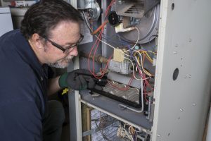 Furnace repair in Ocean County being done by worker in blue overalls and black work gloves shining a flashlight into an open furnace